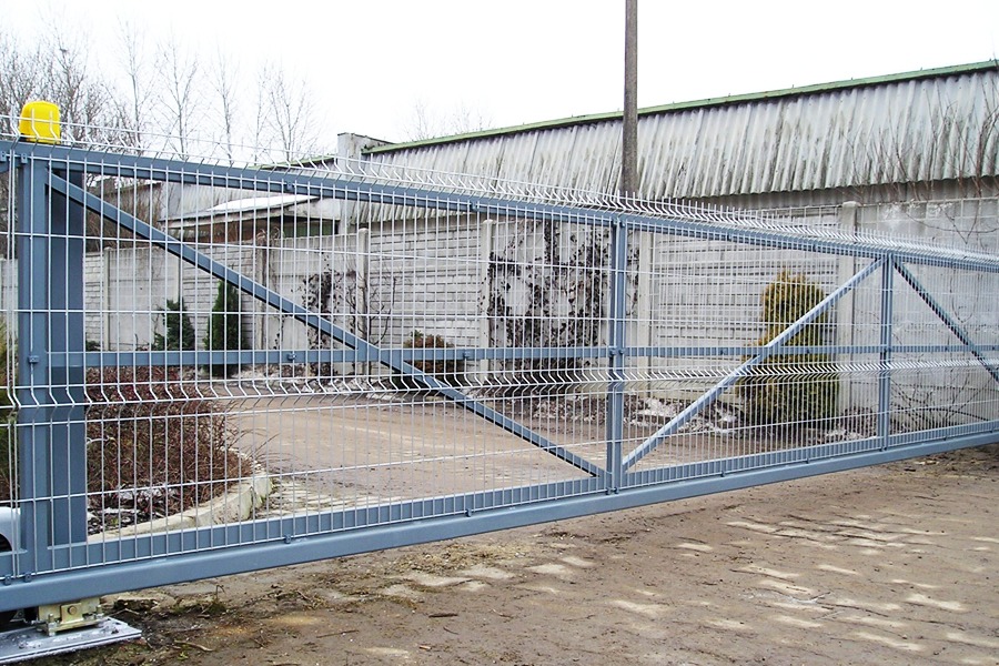 Przesuwna brama metalowa z siatki z żółtą lampą ostrzegawczą, widoczna na tle betonowego ogrodzenia i budynku przemysłowego.