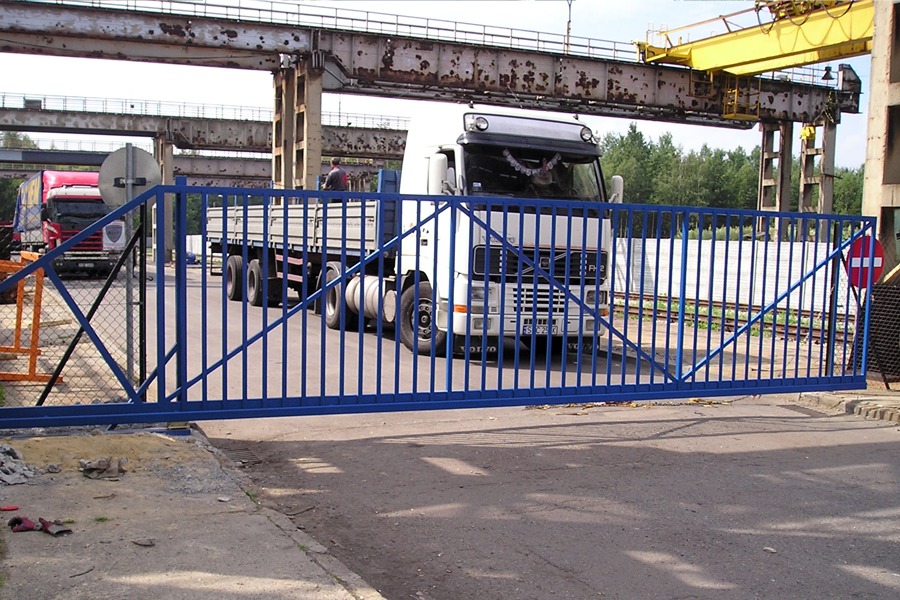 Niebieska, przesuwna brama metalowa na tle ciężarówki i konstrukcji stalowej z suwnicą.