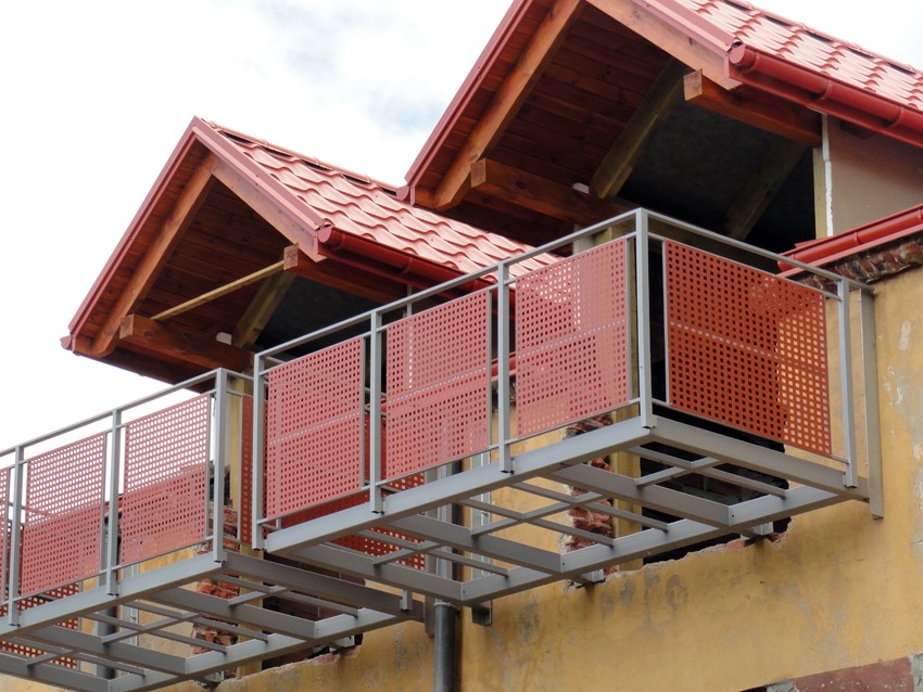 Dwa balkony z metalową konstrukcją i czerwonymi, perforowanymi panelami na tle budynku z czerwonym dachem, widoczne fragmenty muru pod konstrukcją balkonu.