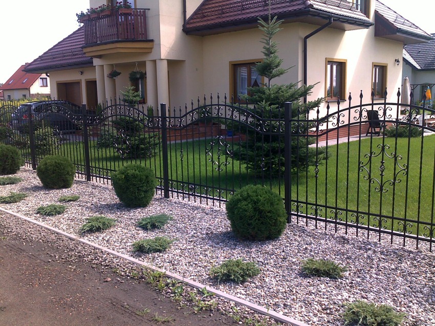Czarny, metalowy płot z ozdobnymi elementami, oddzielający posesję z zadbanym trawnikiem i krzewami, widok na dom z balkonem i zaparkowany samochód.