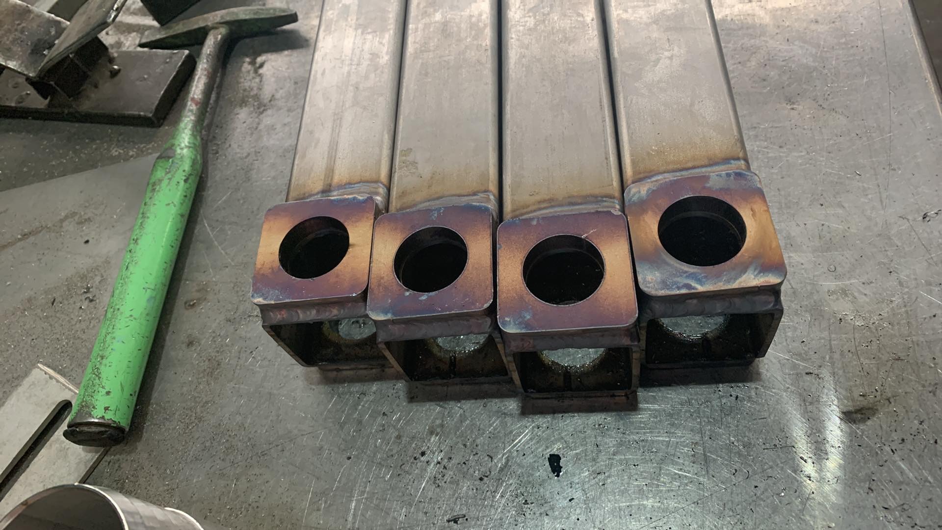 Cztery metalowe profile spawane z okrągłymi otworami, leżące na metalowym stole warsztatowym obok zielonego młotka, widoczne ślady spawania.