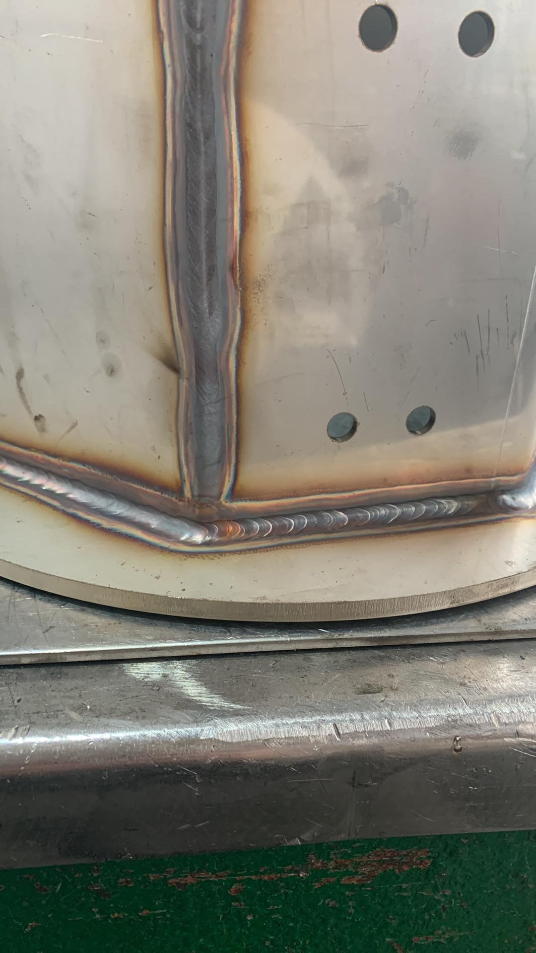 Szczegółowe zbliżenie spoiny na metalowym zbiorniku z widocznymi otworami, wykonanej metodą spawania łukowego, z charakterystycznymi przebarwieniami termicznymi.