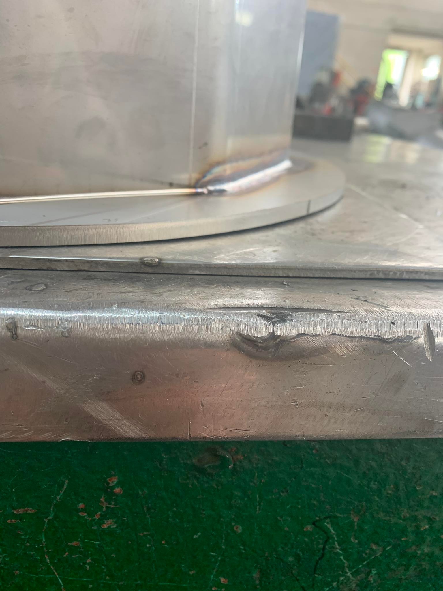 Metalowy element po spawaniu, widoczne ślady obróbki i spawania, tło warsztatowe z elementami wyposażenia.