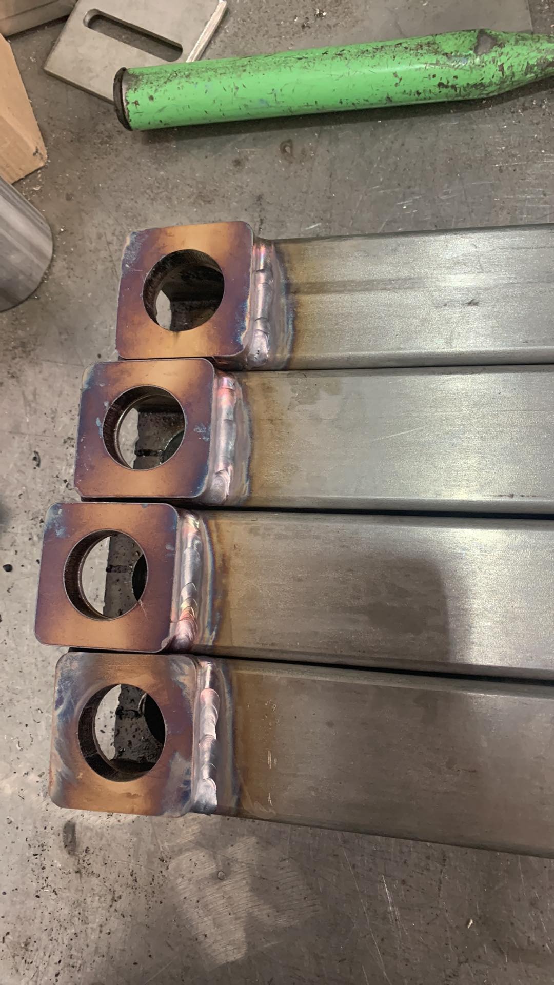 Cztery metalowe profile kwadratowe z przyspawanymi kwadratowymi elementami z otworem, widoczne ślady spawania i przebarwienia termiczne.