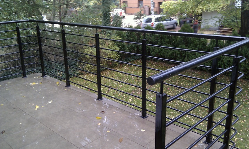 Barierka balkonowa z czarnych, metalowych prętów o minimalistycznym designie, widok z góry na mokrą, wykafelkowaną podłogę balkonu i ogród z drzewami w tle.