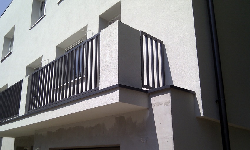 Nowoczesne, czarne ogrodzenie metalowe balkonu w nowym budynku z jasną elewacją, widoczne z dołu pod kątem, z fragmentami niezakończonych prac wykończeniowych.