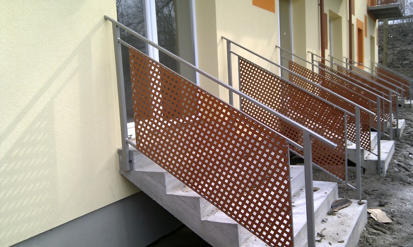 Zewnętrzne schody z ażurową, metalową balustradą w kolorze rdzy, zamontowane przy budynku o jasnej elewacji.