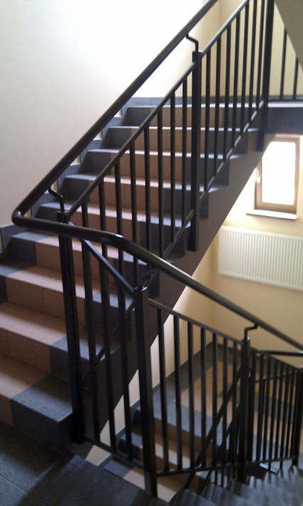 Klatka schodowa w budynku mieszkalnym z metalową balustradą pomalowaną na ciemny kolor, widok z dołu na górę, schody wyłożone płytkami w dwóch odcieniach szarości i beżu.