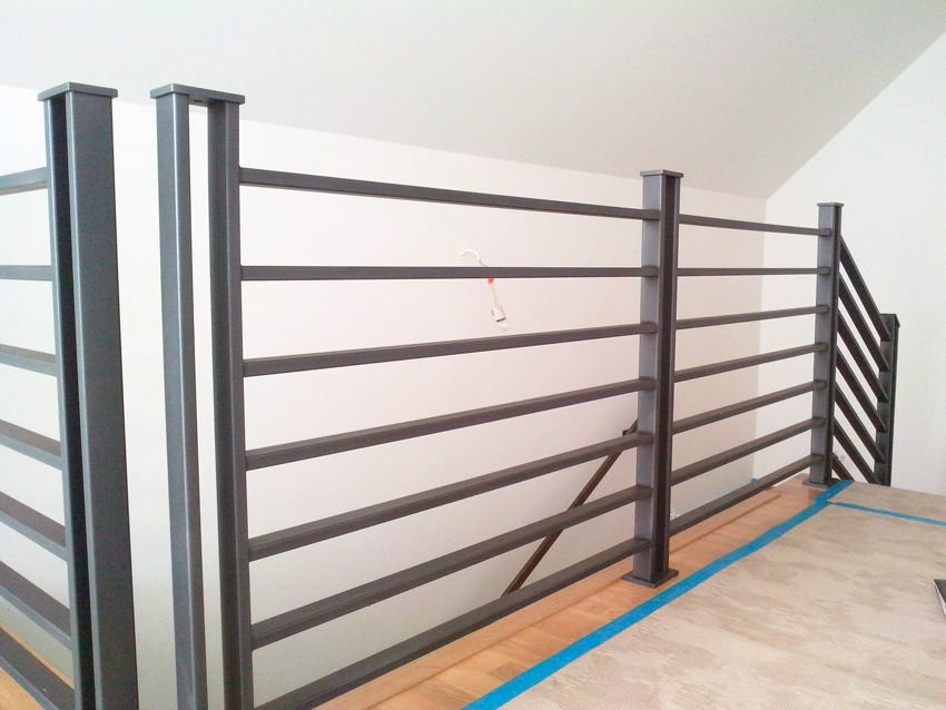 Nowoczesne metalowe balustrady schodowe w kolorze grafitowym z poziomymi szczeblami, widoczne zabezpieczenia podłogi taśmą malarską.