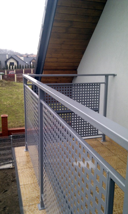 Szare metalowe ogrodzenie balkonowe z perforowanymi panelami w kształcie rombów, widok z dołu, w tle domy jednorodzinne i pochmurne niebo.