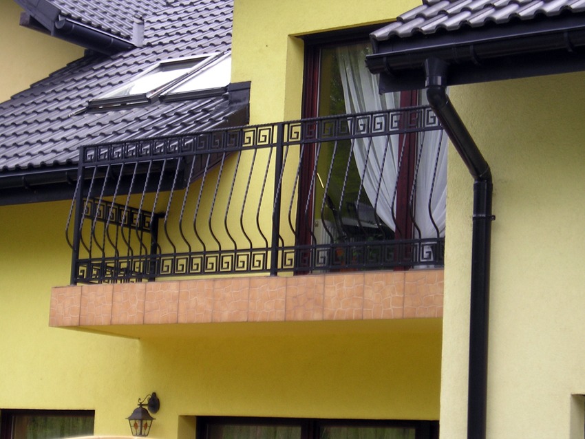 Metalowa balustrada balkonowa z motywem meandru greckiego, zamontowana na balkonie wykończonym płytkami ceramicznymi, na tle żółtej elewacji budynku z widocznymi oknami i dachem z dachówką ceramiczną.