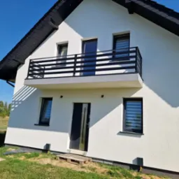 Biały dom z czarnym dachem i balkonem z czarną, poziomą balustradą. Widok z dołu, podkreślający nowoczesny styl i symetrię elewacji.