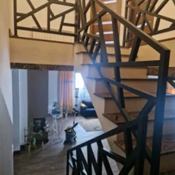 Nowoczesne schody z marmurowymi stopniami i geometryczną, metalową balustradą w ciemnym kolorze. Widok z góry, wnętrze domu z roślinami i odkurzaczem w tle.