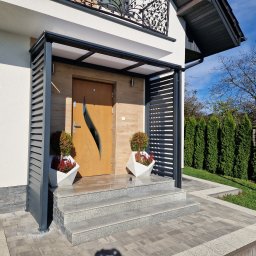 Stalowi Bracia - Nowoczesna zabudowa wejścia do domu z szarej metalowej konstrukcji z poziomymi lamelami, drewniane drzwi z dekoracyjnym wycięciem, schody z jasnego kamienia.