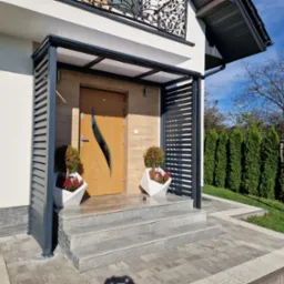 Nowoczesna zabudowa wejścia do domu z szarej metalowej konstrukcji z poziomymi lamelami, drewniane drzwi z dekoracyjnym wycięciem, schody z jasnego kamienia.