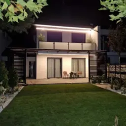 Nowoczesna, podświetlana pergola przy białym domu z balkonem. Minimalistyczny design, zadbany trawnik i dekoracyjne kamienie. Oświetlenie LED podkreśla architekturę.