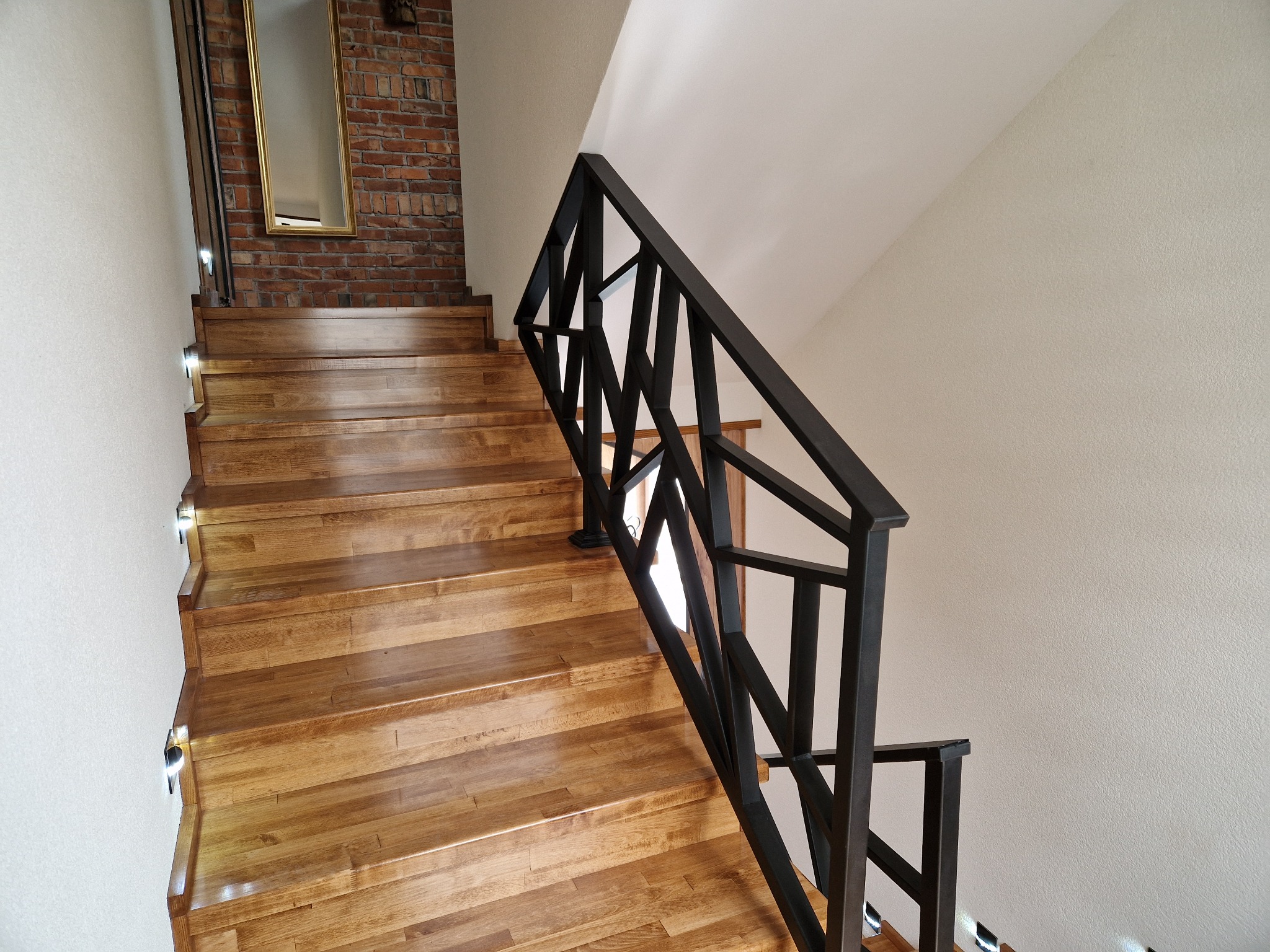 Drewniane schody z czarną, geometryczną balustradą. Na ścianie cegła i lustro. Oświetlenie punktowe na stopniach. Nowoczesny design z elementami rustykalnymi.
