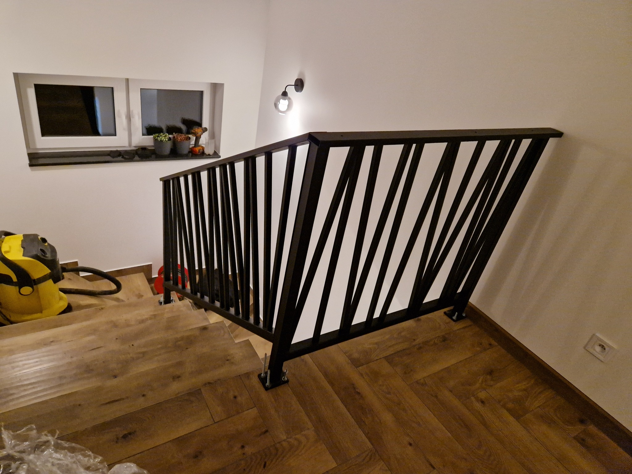 Nowoczesna, czarna barierka schodowa z ukośnymi prętami, zamontowana na drewnianych schodach. U dołu widoczny odkurzacz, a w tle okno z roślinami.