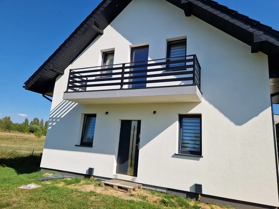 Biały dom z czarnym dachem i balkonem z czarną, poziomą balustradą. Widok z dołu, podkreślający nowoczesny styl i symetrię elewacji.
