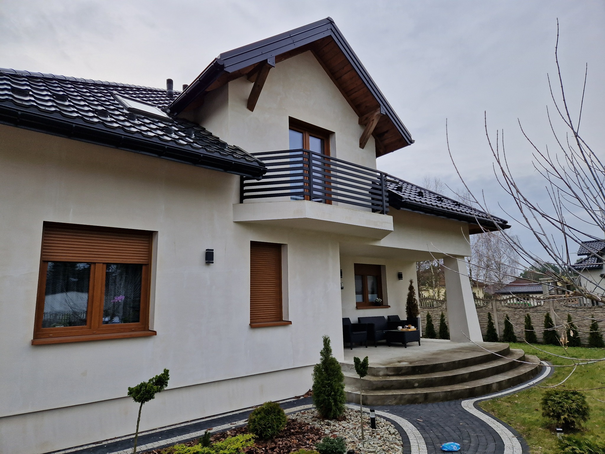 Dwukondygnacyjny dom z balkonem z czarną, poziomą konstrukcją. Widoczne okna z brązowymi ramami, czarny dach i fragment ogrodu z ozdobnymi krzewami.