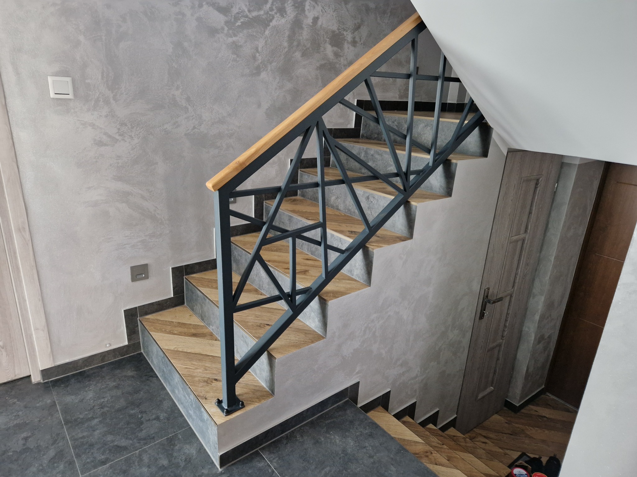 Schody z drewnianymi stopniami i metalową, geometryczną konstrukcją. Szara ściana z efektem dekoracyjnym w tle. Nowoczesne wykończenie wnętrza.