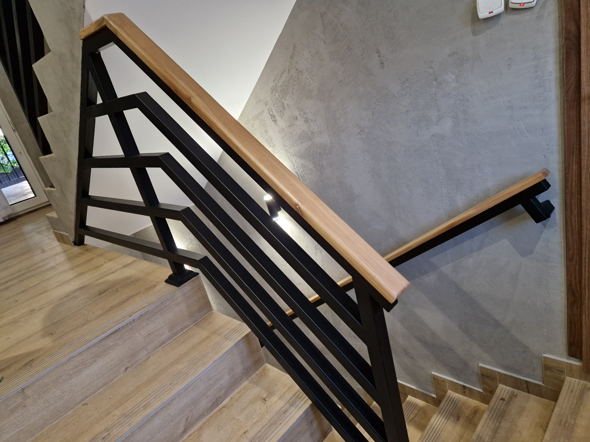 Nowoczesne schody z drewnianymi stopniami i czarną, geometryczną balustradą z drewnianą poręczą. Widok z góry, podkreślający detale wykończenia ścian.