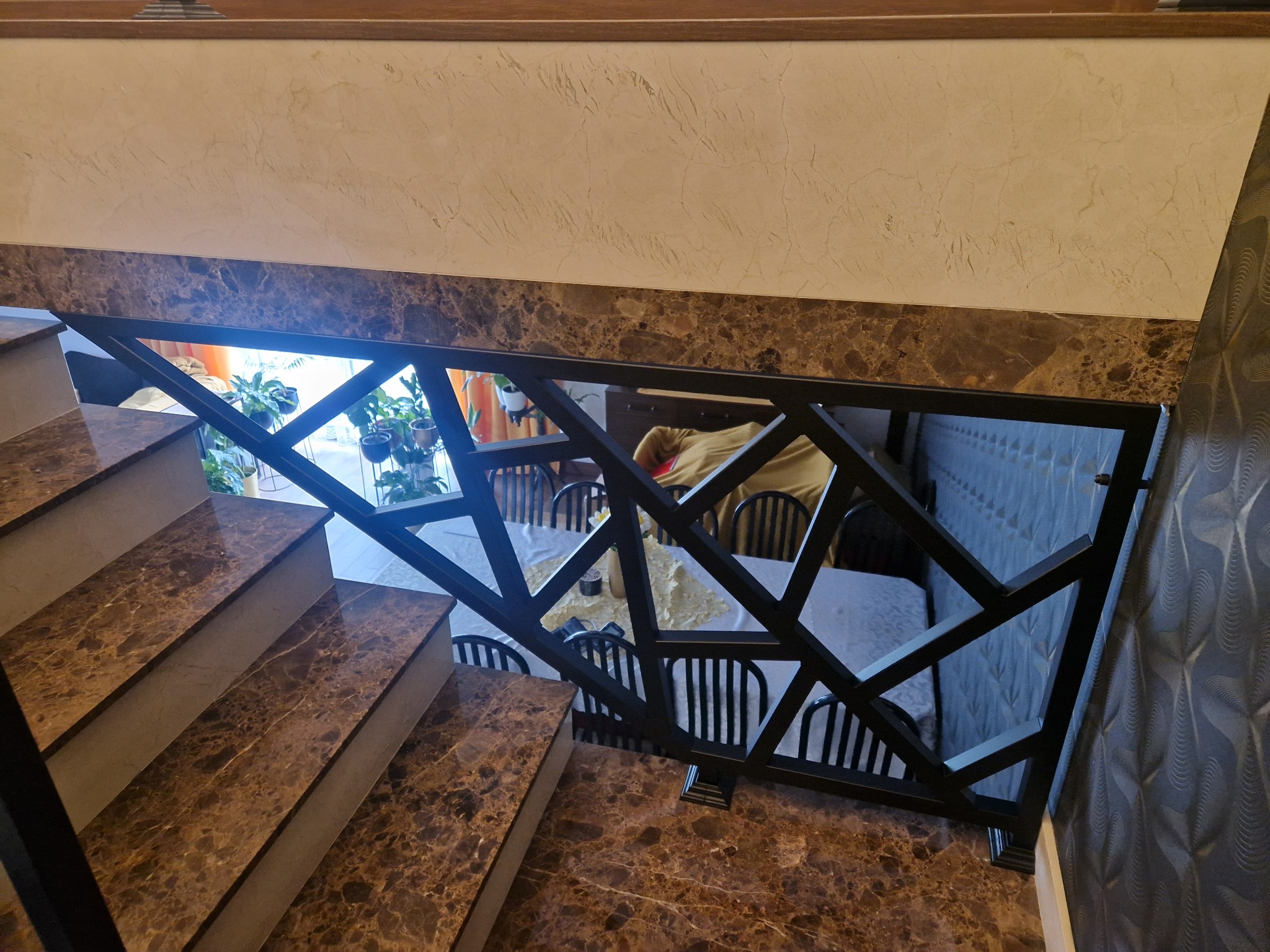 Schody z marmurowymi stopniami i czarną, geometryczną balustradą metalową. W tle widoczny stół i krzesła w pokoju.