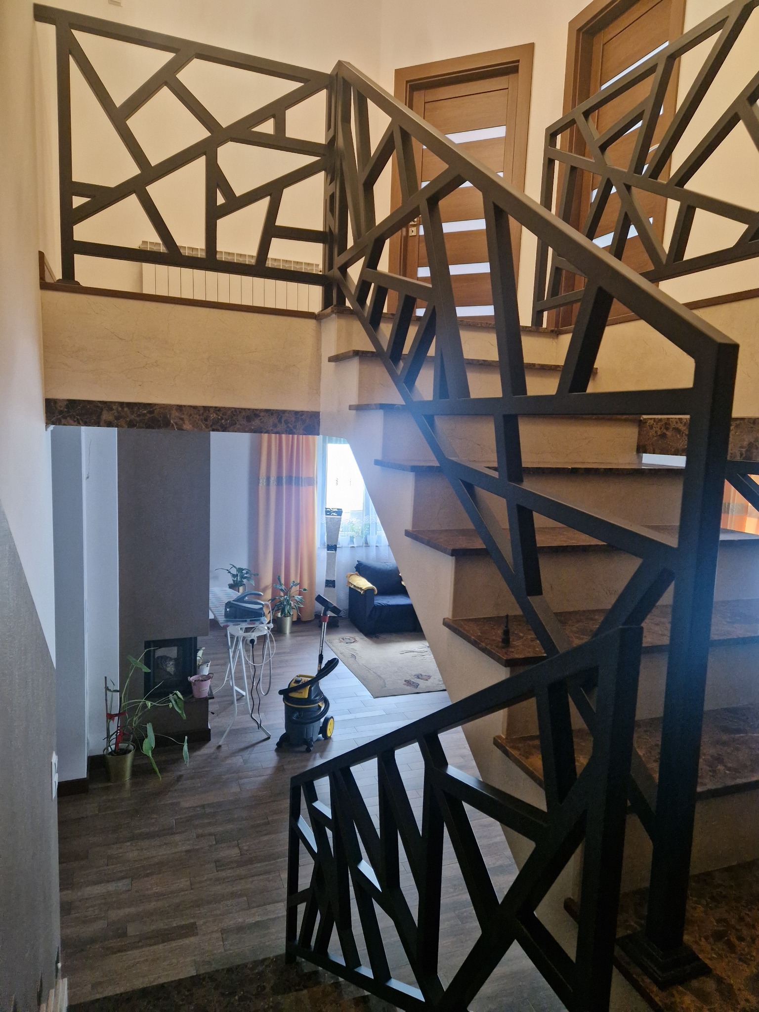 Nowoczesne schody z marmurowymi stopniami i geometryczną, metalową balustradą w ciemnym kolorze. Widok z góry, wnętrze domu z roślinami i odkurzaczem w tle.