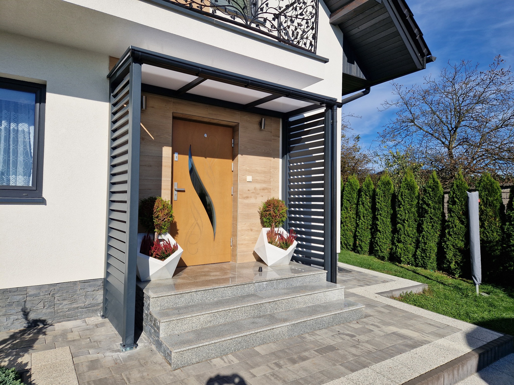 Nowoczesna zabudowa wejścia do domu z szarej metalowej konstrukcji z poziomymi lamelami, drewniane drzwi z dekoracyjnym wycięciem, schody z jasnego kamienia.
