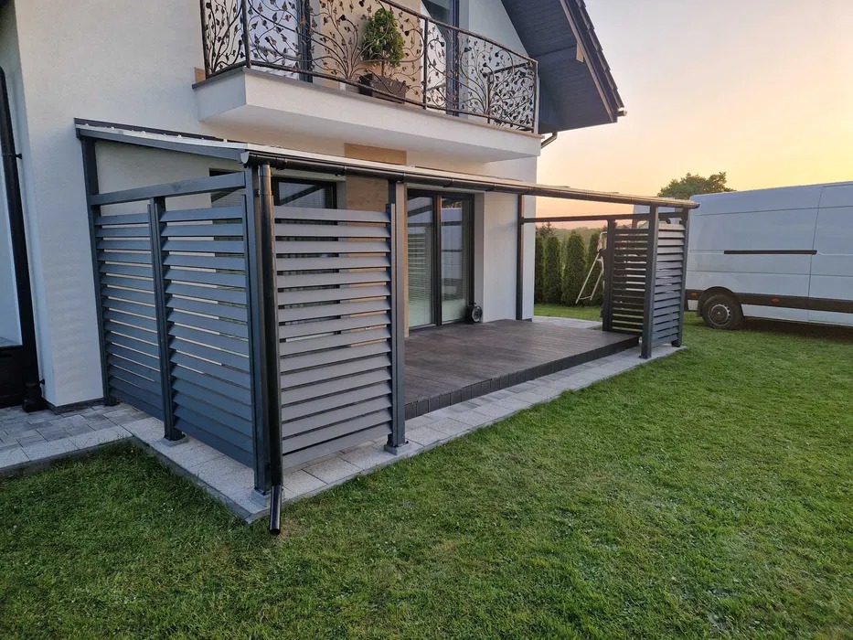 Nowoczesna, szara pergola z poziomymi lamelami, przylegająca do domu z balkonem z dekoracyjną, metalową balustradą. Widoczny fragment trawnika i zaparkowany biały bus.