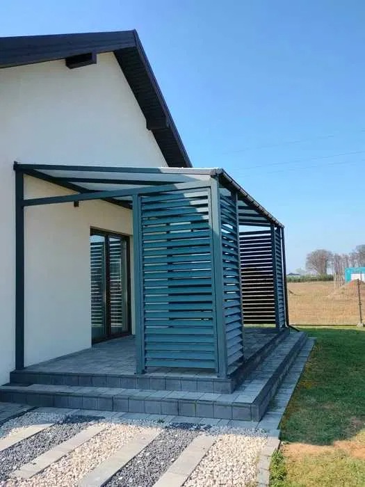 Nowoczesna pergola metalowa z lamelami, tworząca zacienione przejście w Zrąbcu. Widoczna konstrukcja i wykończenie w kolorze grafitowym, dopasowane do elewacji budynku.