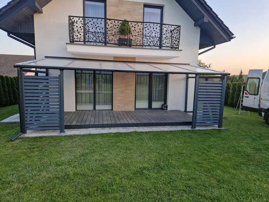 Metalowa pergola z zadaszeniem i ażurowymi ściankami na drewnianym tarasie, z balkonem z dekoracyjną balustradą w tle, na tle domu z ogrodem.