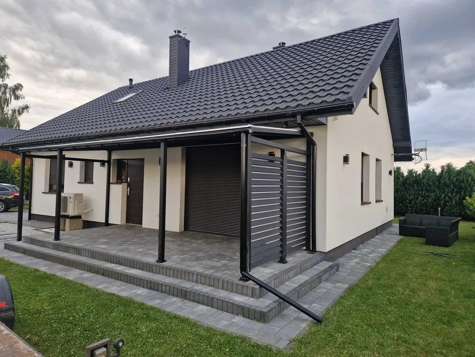 Biały dom z czarnym dachem i pergolą z metalowymi panelami. Widoczne schody z kostki brukowej i trawnik. Całość w nowoczesnym stylu.