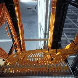 Luminus Katsiaryna Koshaleva - Pomarańczowe kable światłowodowe ułożone w metalowej konstrukcji w hali, z widokiem na podłogę i sufit. Kable spięte czarnymi opaskami. Widok z góry.