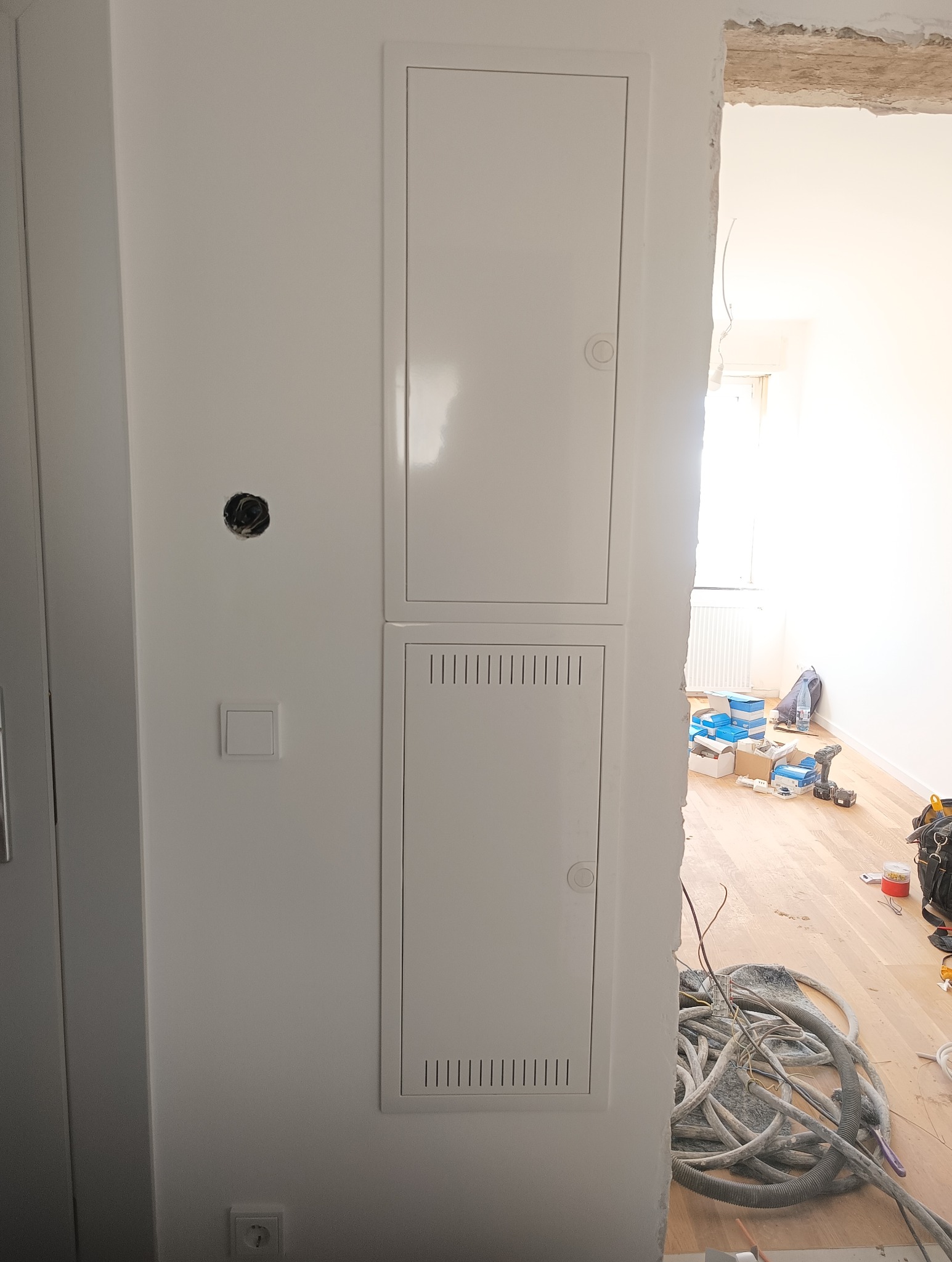 Biała, podwójna skrzynka elektryczna na ścianie w trakcie montażu. Widoczne kable i narzędzia w tle. Otwór na gniazdko i włącznik obok.