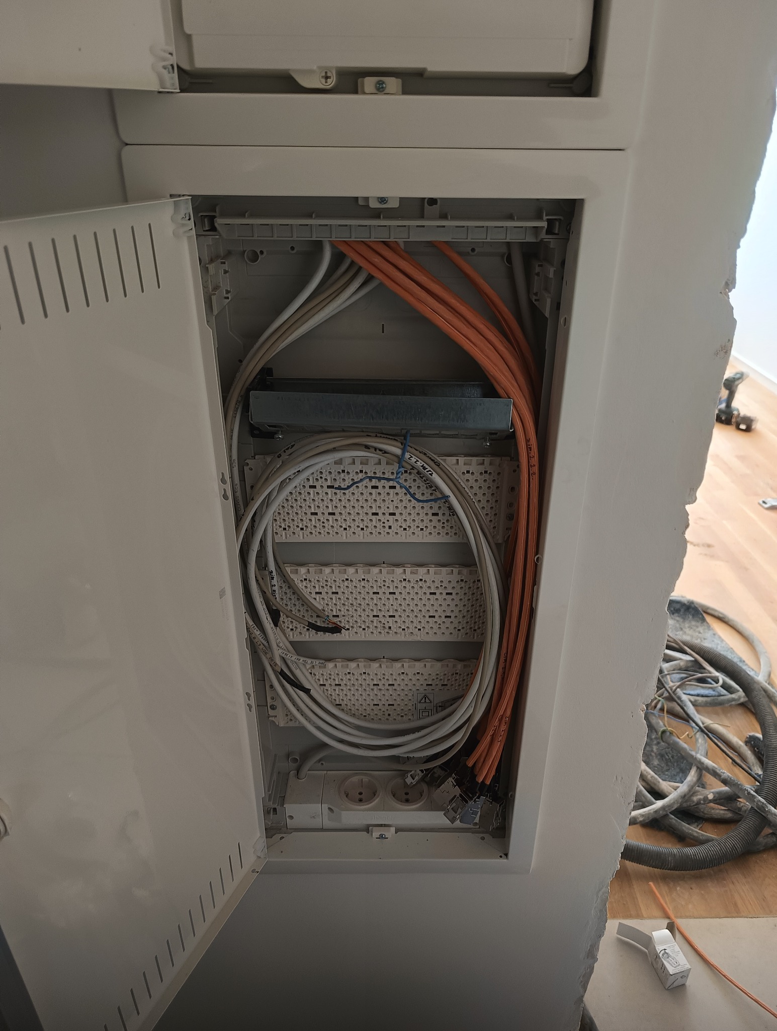 Otwarta skrzynka z przewodami elektrycznymi w ścianie w Skarszewach. Widoczne kable w kolorze białym i pomarańczowym. Instalacja w trakcie prac, widoczny bałagan.