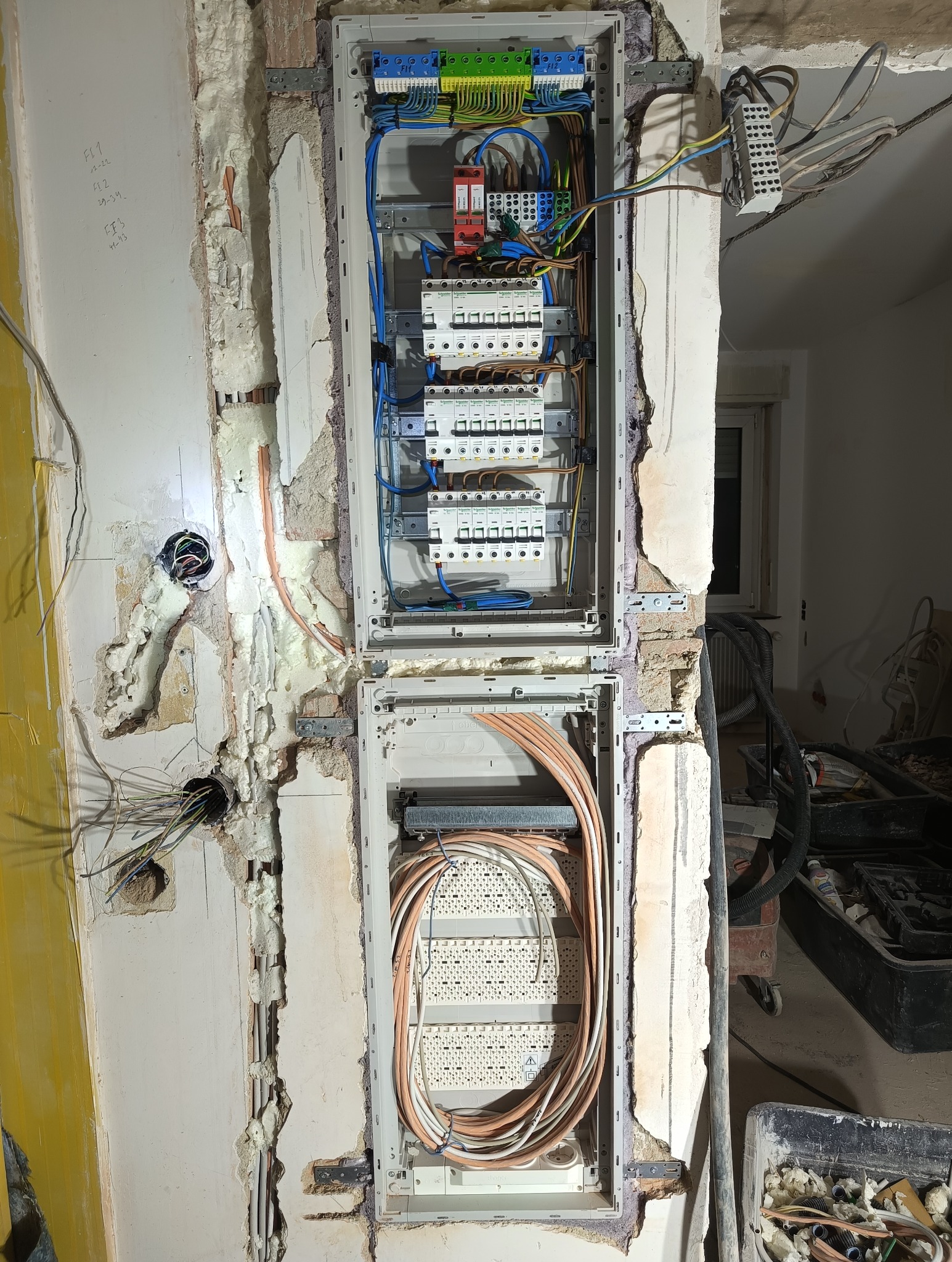 Dwu-częściowa skrzynka elektryczna w trakcie montażu w ścianie, widoczne przewody i elementy instalacji. Ściana w stanie surowym, z widocznymi śladami prac.