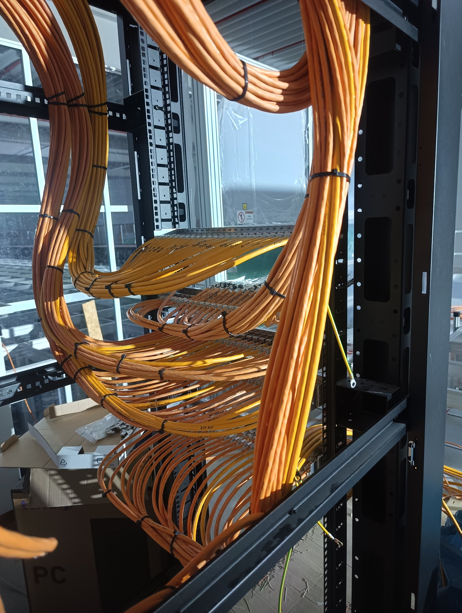 Pomarańczowe kable elektryczne ułożone w szafie rack, zabezpieczone czarnymi opaskami. Widoczne złącza i okablowanie w tle, częściowo poza kadrem.