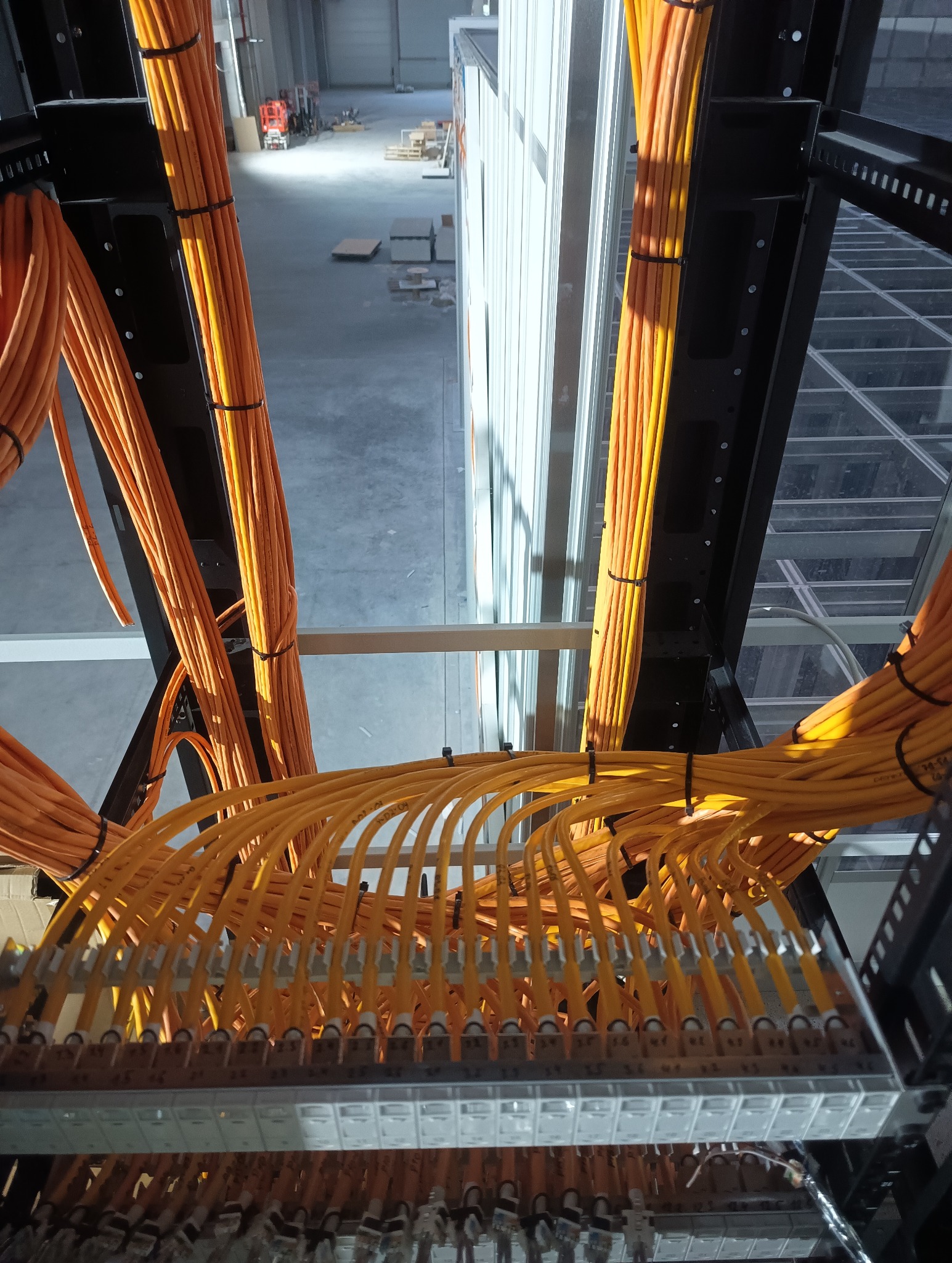 Pomarańczowe kable światłowodowe ułożone w metalowej konstrukcji w hali, z widokiem na podłogę i sufit. Kable spięte czarnymi opaskami. Widok z góry.