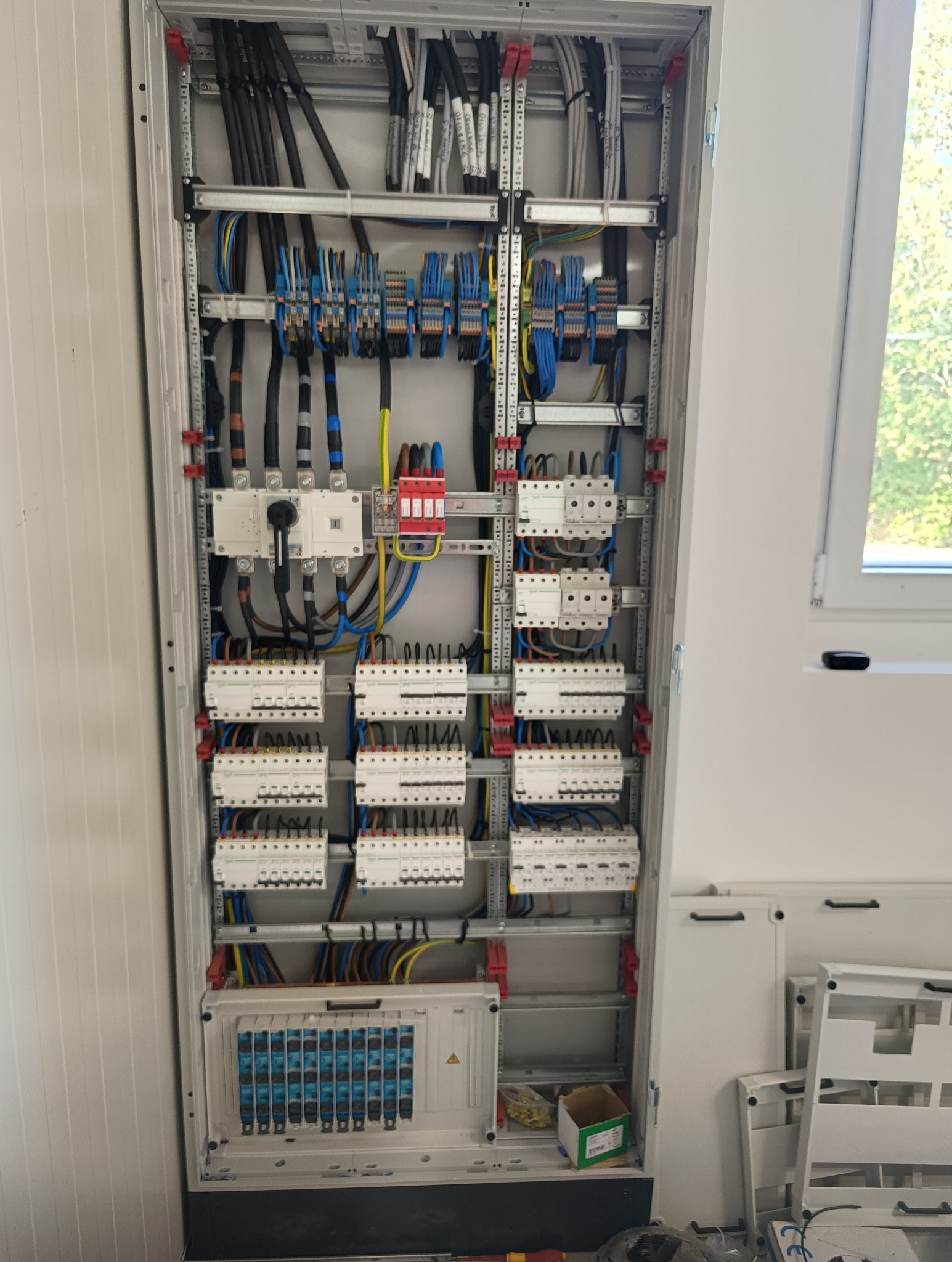 Rozdzielnica elektryczna w metalowej obudowie z licznymi zabezpieczeniami, przewodami i elementami montażowymi. Widoczne okno w tle.