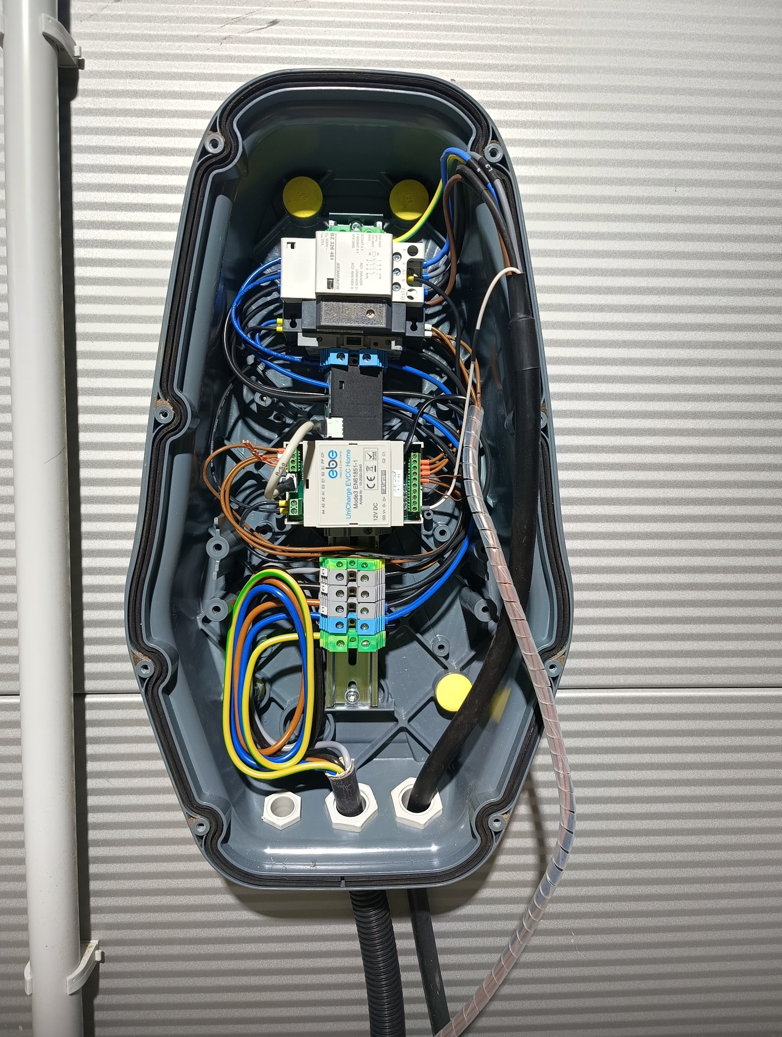 Szara skrzynka z podłączonymi przewodami elektrycznymi, widoczne elementy sterujące i złącza. Instalacja elektryczna wewnątrz obudowy na tle szarej ściany.