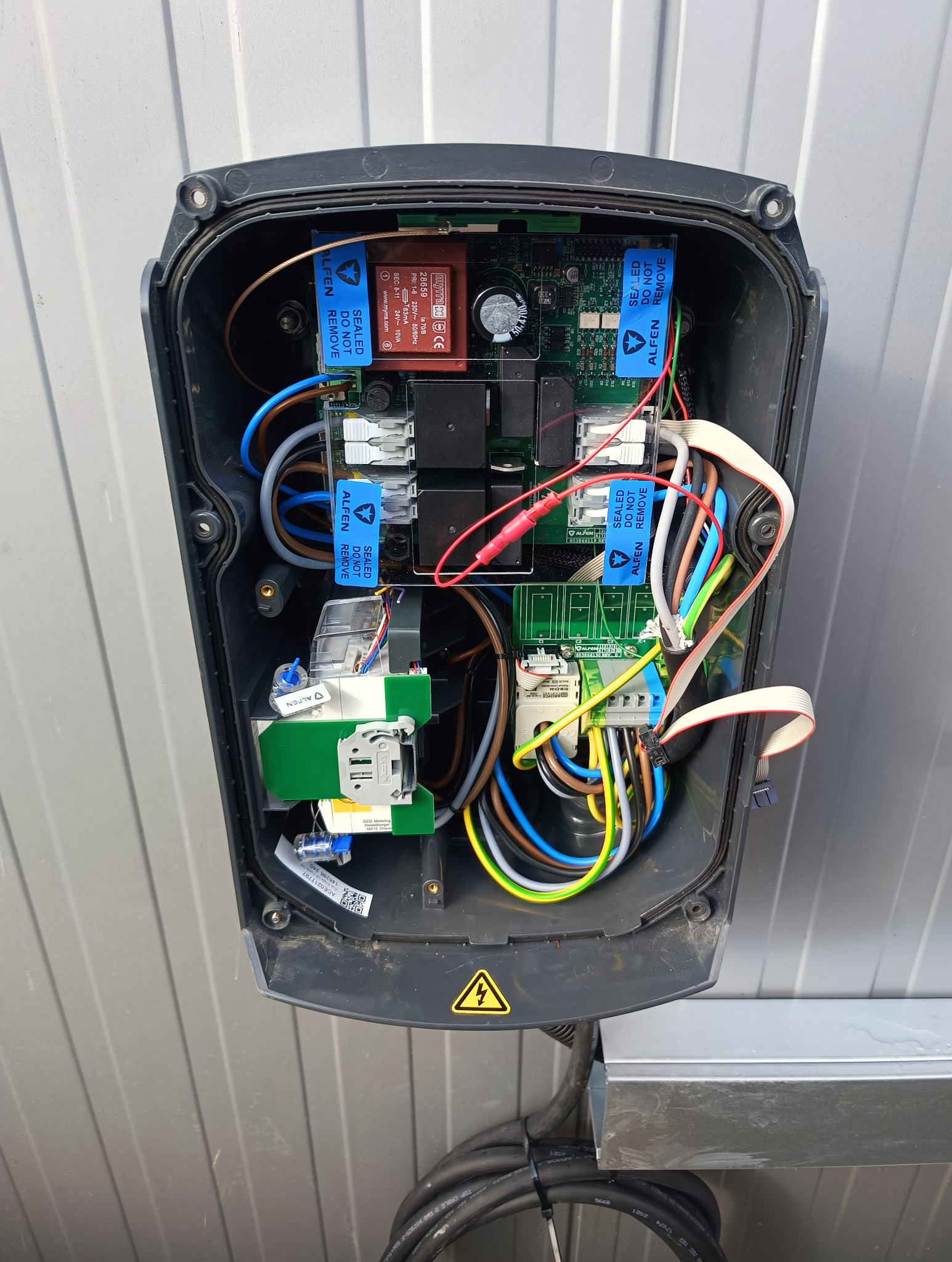 Otwarta obudowa stacji ładowania pojazdów elektrycznych Alfen z widocznymi komponentami elektrycznymi, przewodami i naklejkami ostrzegawczymi. Widok z bliska na wnętrze urządzenia.