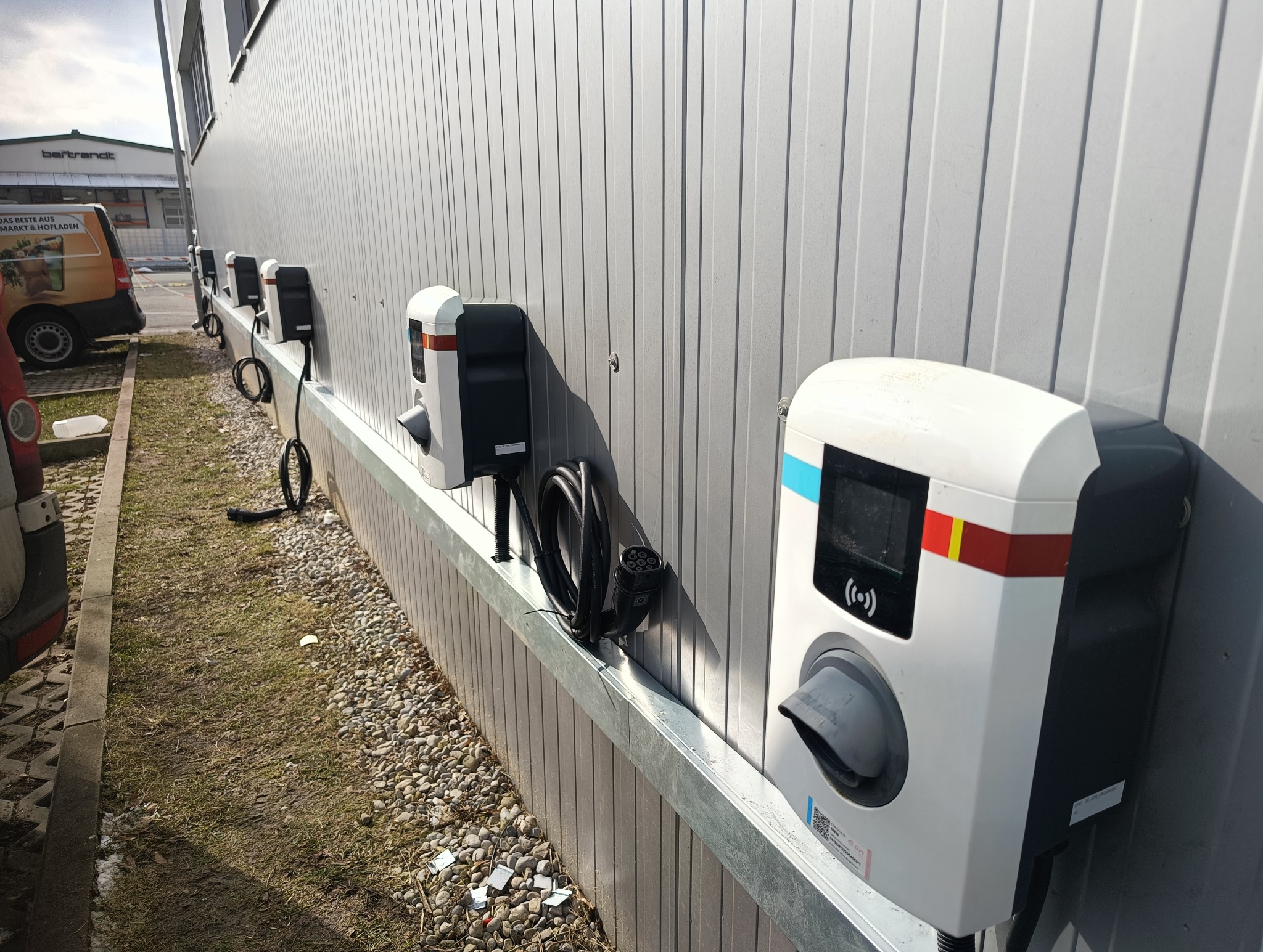 Rząd zamontowanych na ścianie stacji ładowania pojazdów elektrycznych z kablami, widok z boku, w tle budynek. Strefa ładowania aut.