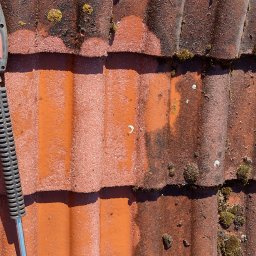 Łukasz Gibas - Częściowo oczyszczona, czerwona dachówka karpiówka z widocznymi śladami mchu i porostów, z użyciem myjki ciśnieniowej, na której widać fragment lancy.