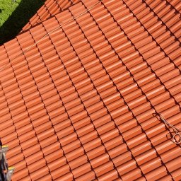 Łukasz Gibas - Nowy, pomarańczowy dach z dachówki ceramicznej, widok z góry, drabina w lewym dolnym rogu, sąsiedni budynek i zielona roślinność w tle.