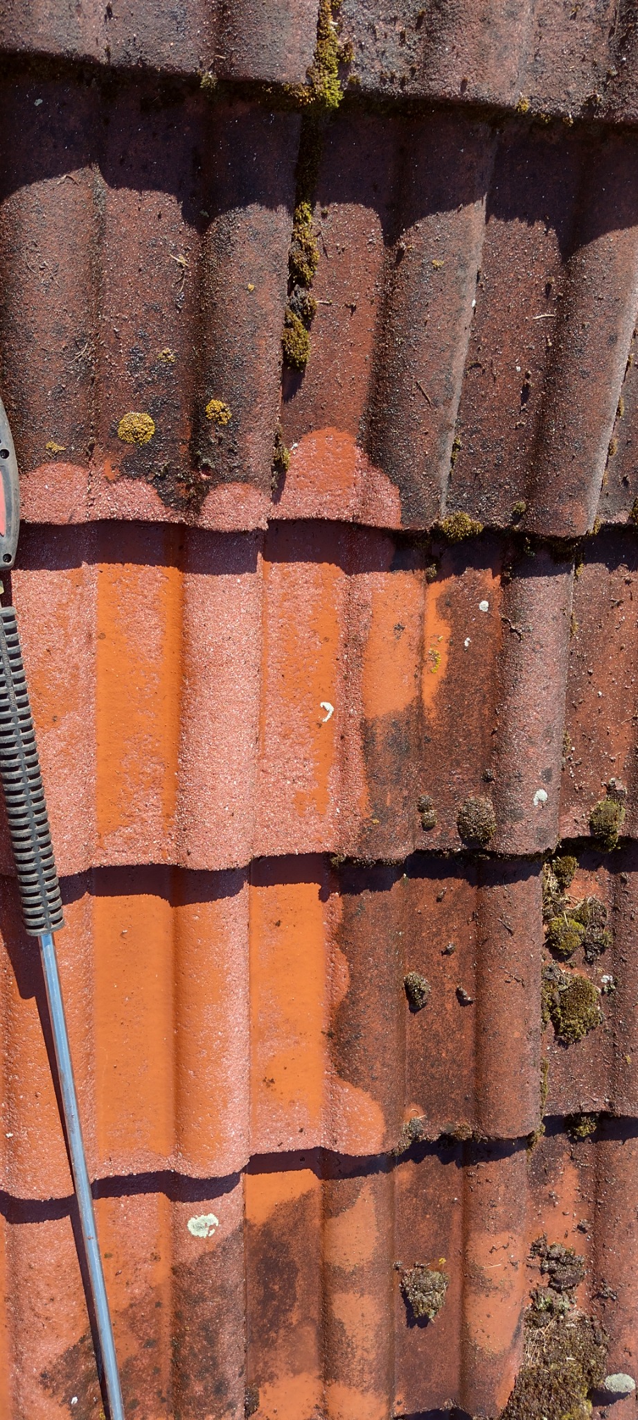 Częściowo oczyszczona, czerwona dachówka karpiówka z widocznymi śladami mchu i porostów, z użyciem myjki ciśnieniowej, na której widać fragment lancy.