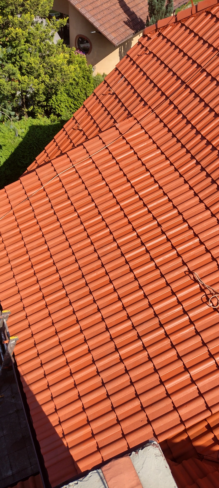 Nowy, pomarańczowy dach z dachówki ceramicznej, widok z góry, drabina w lewym dolnym rogu, sąsiedni budynek i zielona roślinność w tle.