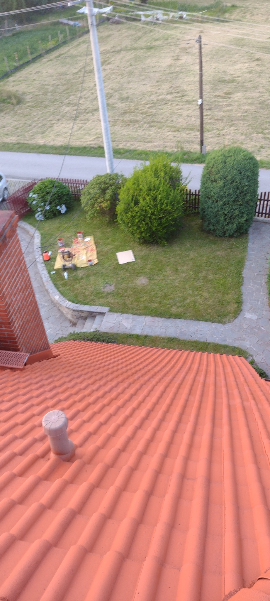 Widok z góry na pomarańczowy dach z kominkiem wentylacyjnym, w tle ogród z rozłożonymi narzędziami malarskimi na żółtej płachcie.