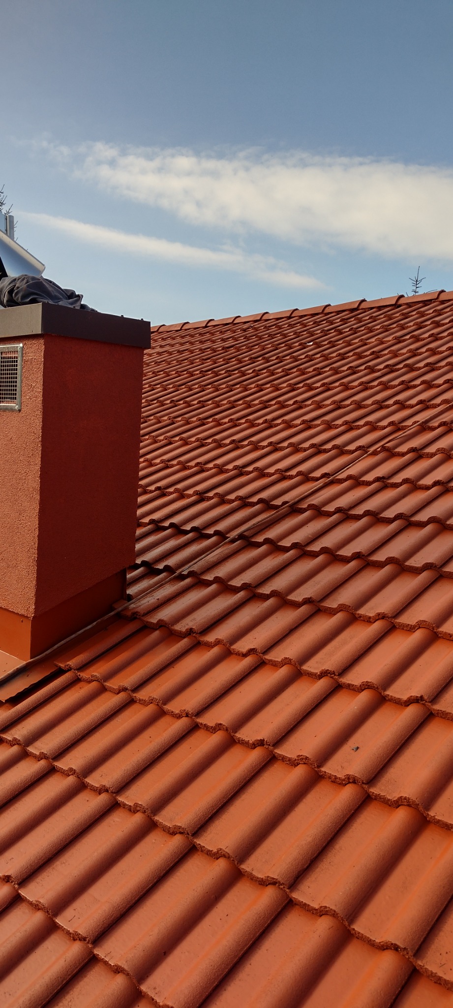 Czerwona dachówka ceramiczna na dachu domu, widoczny komin z kratką wentylacyjną i fragment błękitnego nieba z chmurami.