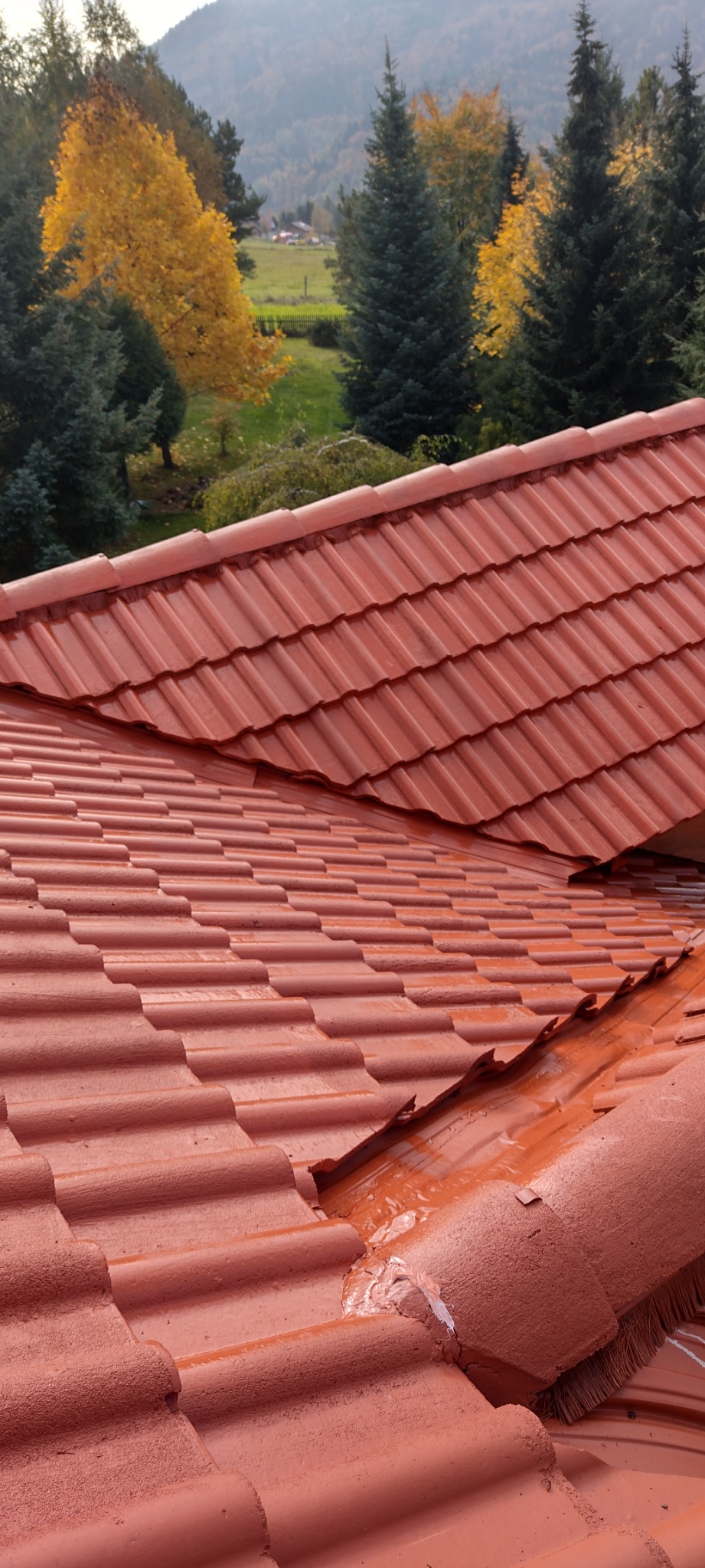 Czerwona dachówka ceramiczna na tle zielonych drzew i wzgórz, widoczne ślady naprawy z użyciem masy uszczelniającej, nierównomiernie nałożona farba na krawędzi dachu.
